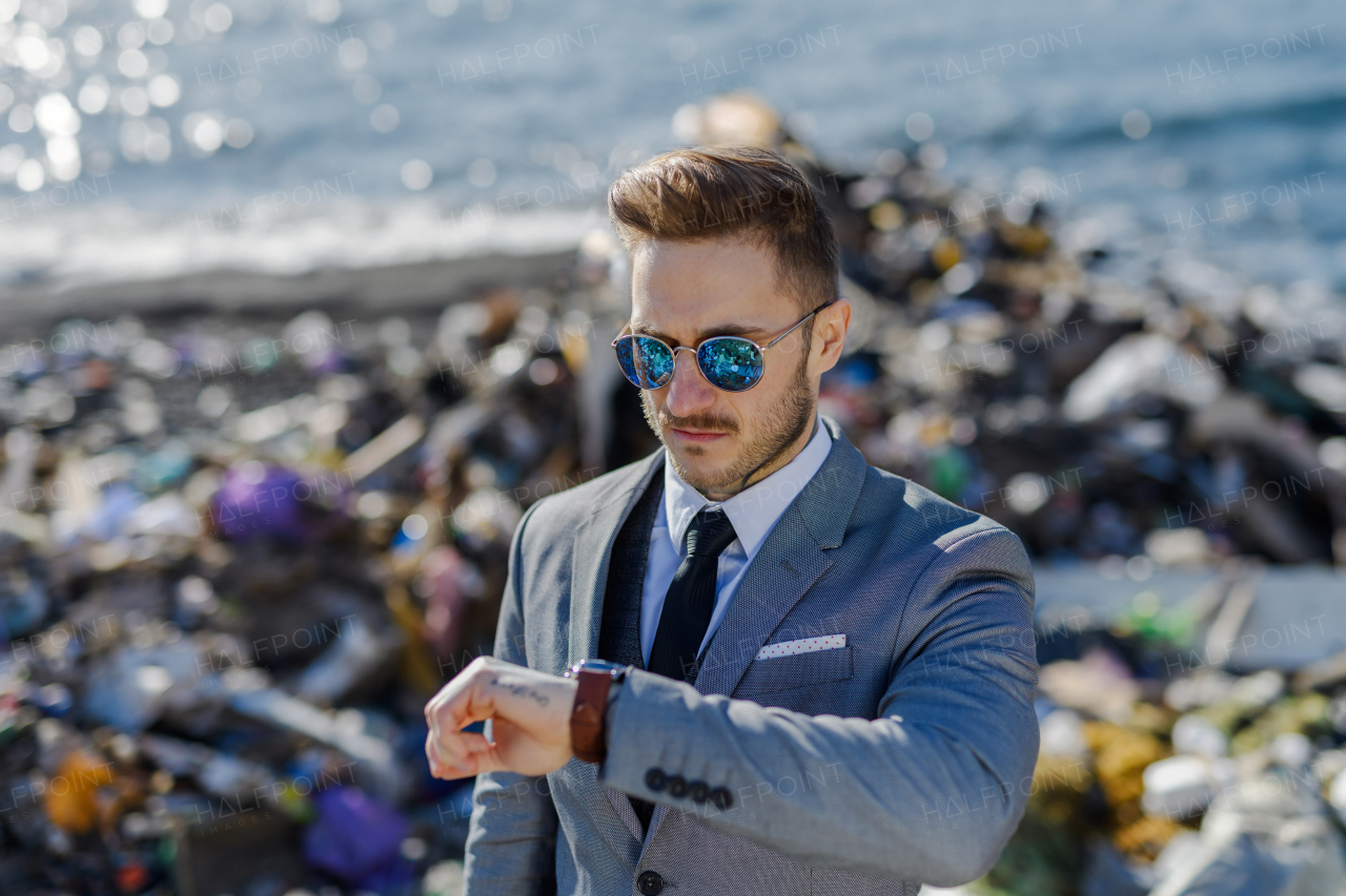 Fashionable modern businessman looking at watch, checking time standing on pile of waste on beach. Consumerism versus pollution concept. Corporate social responsiblity in business.