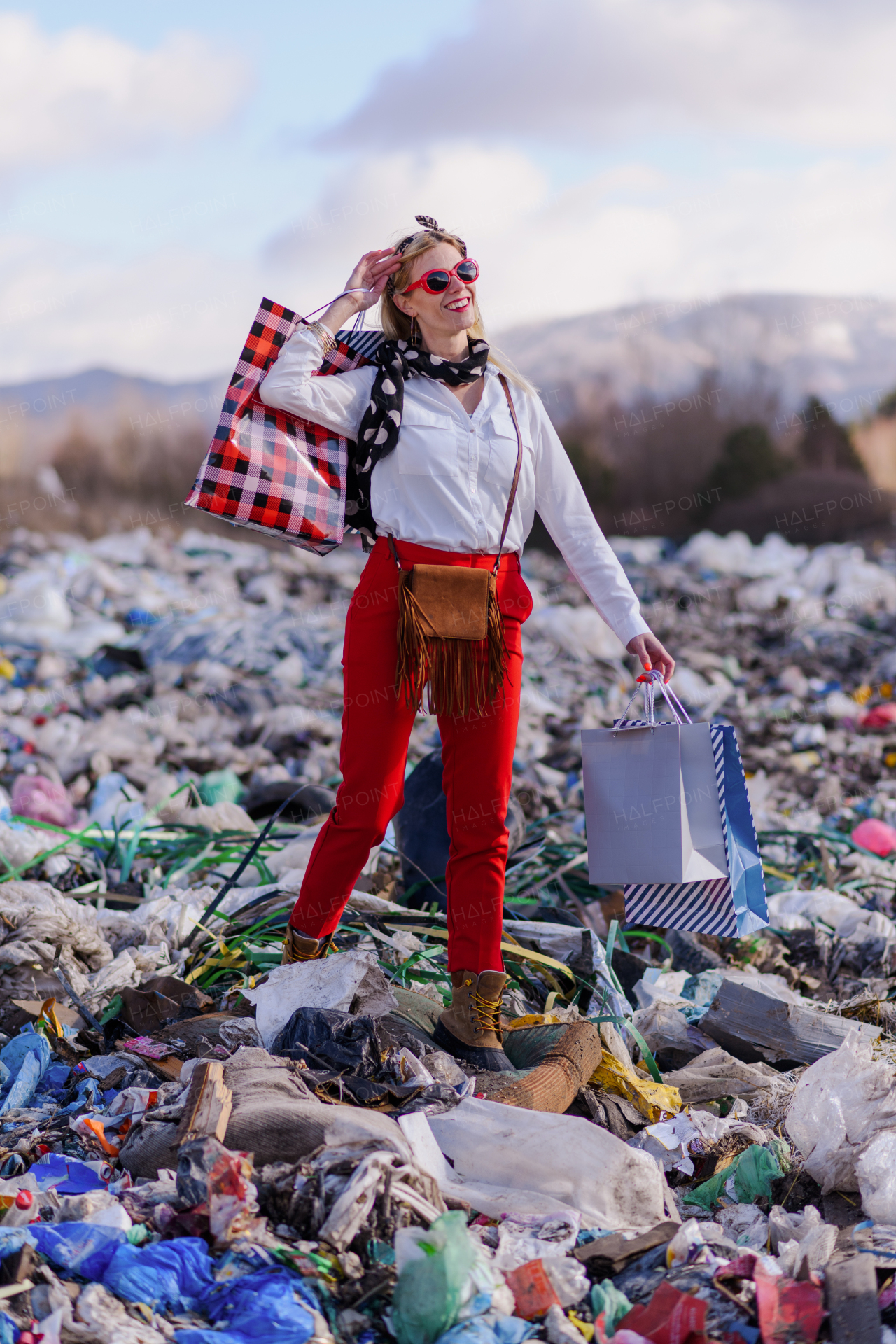 Fashionable modern woman on landfill with shopping bags, consumerism versus pollution concept.