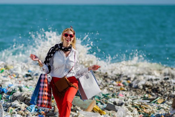 Fashionable modern woman on landfill with shopping bags, standing on pile of waste on beach. Consumerism versus pollution concept.