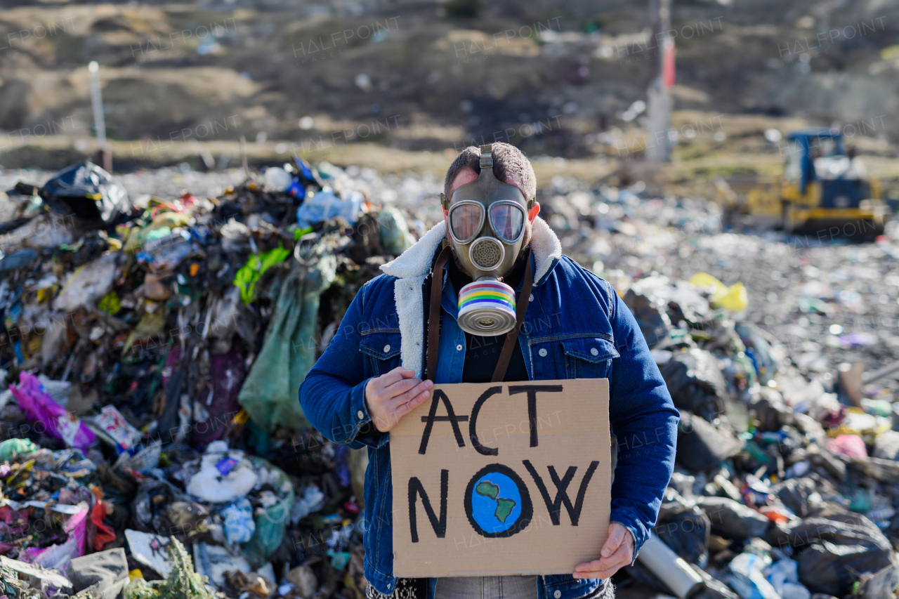 Man activist with gas mask holding placard, protest sign, standing standing on landfill, large pile of waste, environmental concept and eco activism.