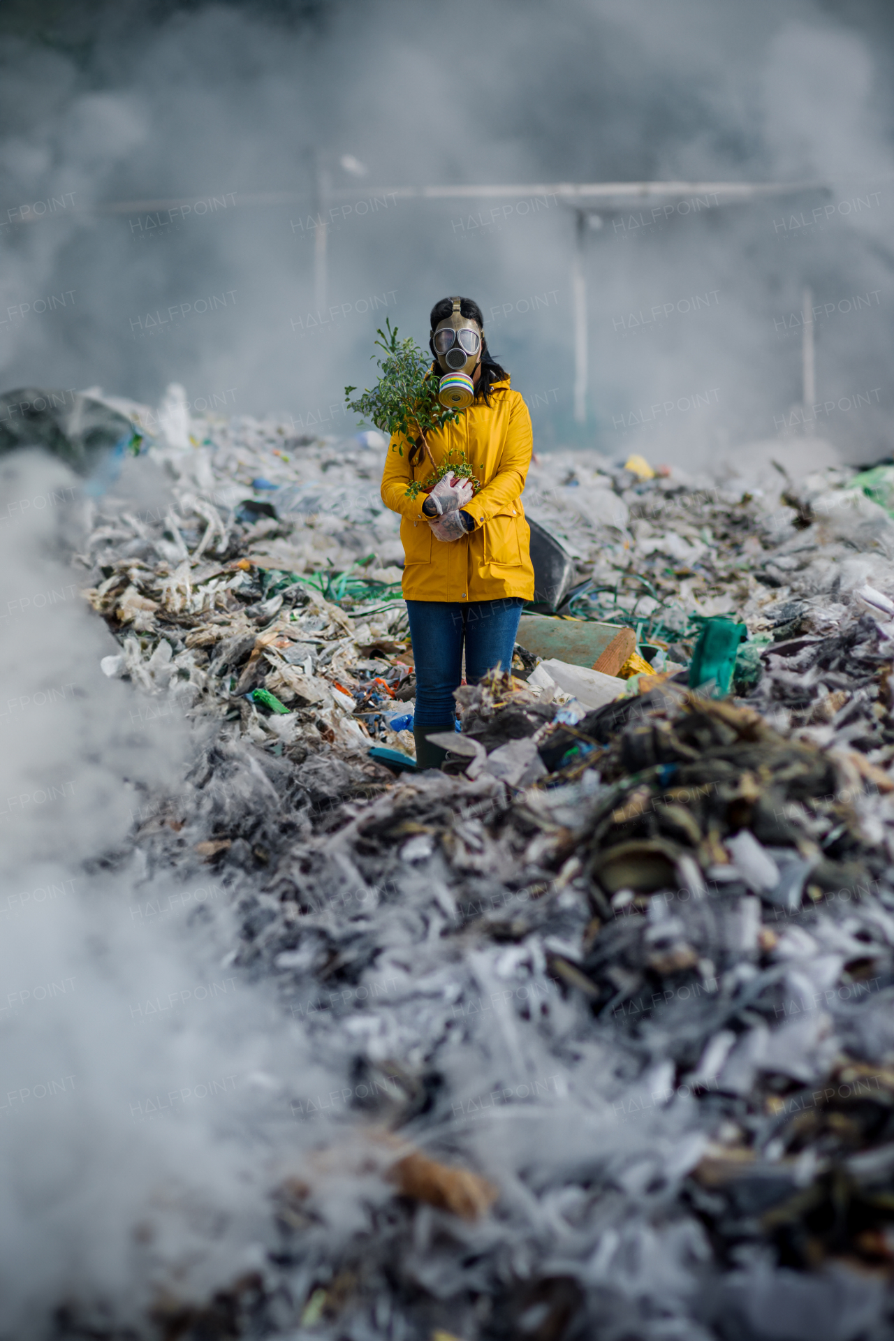 Female activist with gas mask holding potted plant standing on landfill, large pile of waste, environmental concept and eco activism.