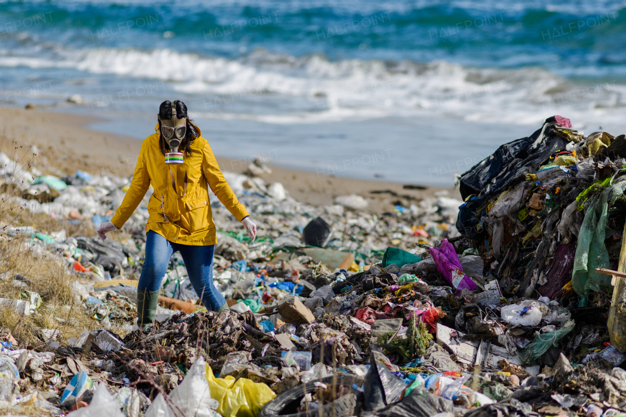 Female activist with gas mask walking on landfill, large pile of waste by sea, shoreline or beach, environmental concept and eco activism.