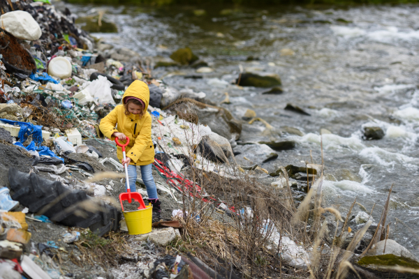 Young girl picking up litter, debris along riverbank in nature. Water and environmental pollution, future generations, eco activism concept.