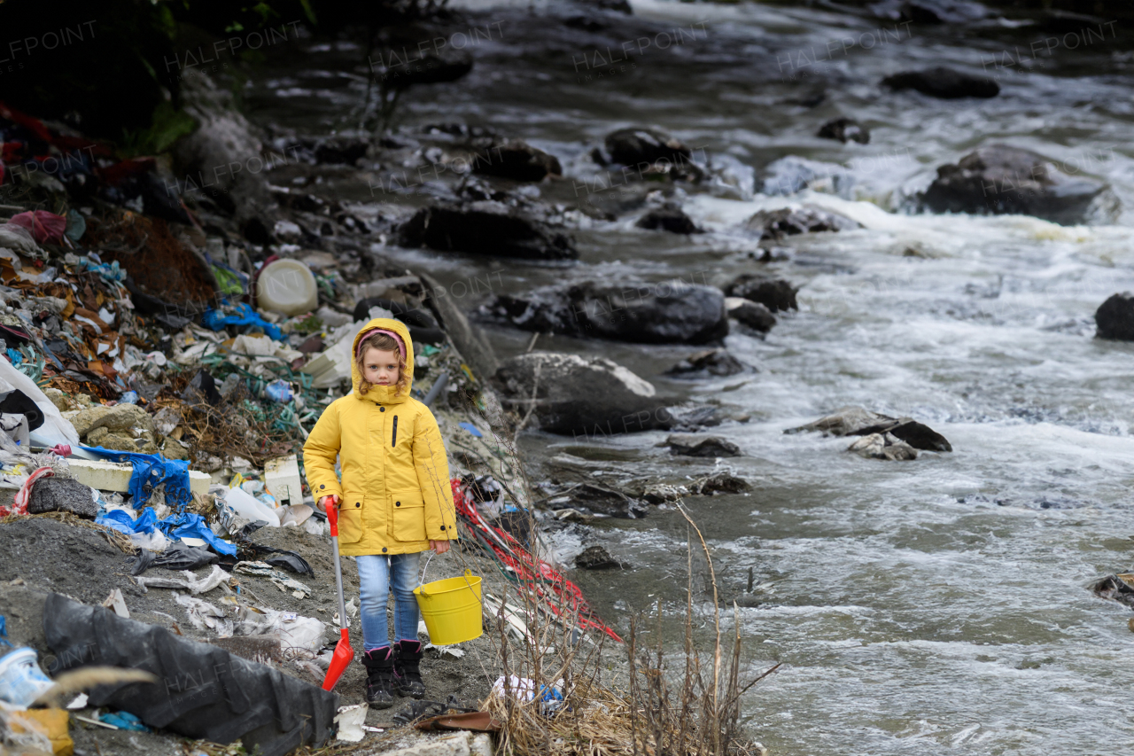 Young girl picking up litter, debris along riverbank in nature. Water and environmental pollution, future generations, eco activism concept.