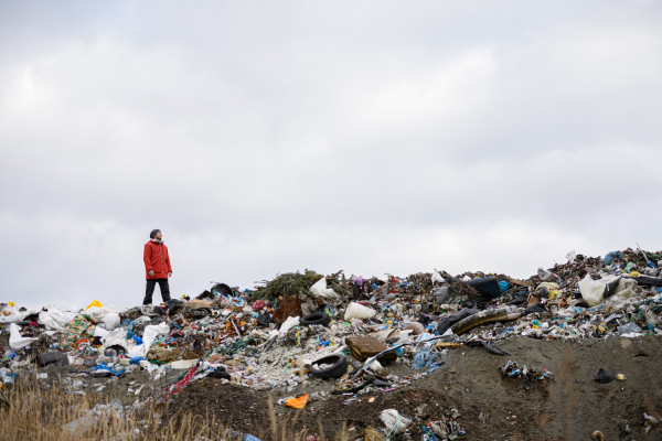 Activist in red jacket standing on landfill, on large pile of waste with dark clouds above. Environmental concept and eco activism.