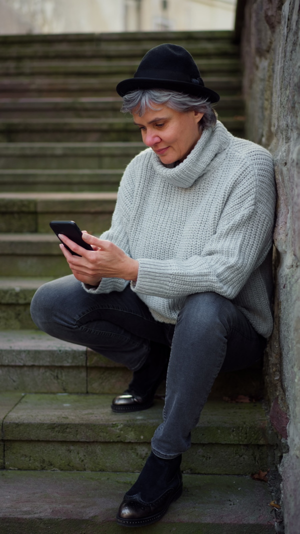 Portrait of mature woman sitting on stairs, using smart phone, smiling and looking into camera. Vertical. Close up.