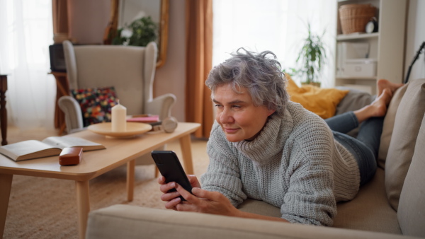 Smiling mature woman using smart phone while lying on sofa at home and relaxing.