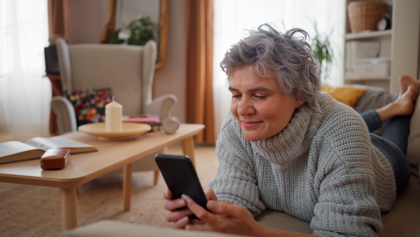 Smiling mature woman using smart phone while lying on sofa at home, looking at camera, smiling. Detail shot.