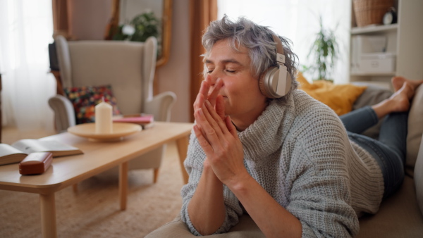 Smiling mature woman listening to music on headphones while lying on sofa at home, smiling. Close up.