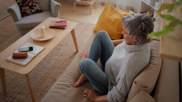 Smiling mature woman putting on headphones and listening to music while sitting on sofa at home. Top view.