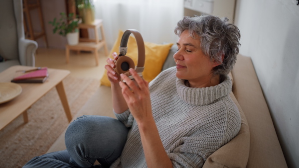 Smiling mature womanputting on headphones and listening to music while sitting on sofa at home, smiling. Close up.