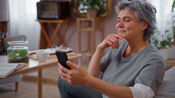 Mature woman sitting on floor in living room at home, using smart phone, smiling. Close up.