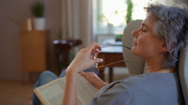 Mature woman reading a book in armchair at home, contemplating, putting on glasses. Detail shot.