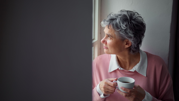 Attractive mature woman looking out of window, drinking coffee. Close up.