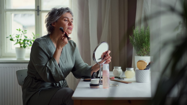 Attractive mature woman at home, sitting at cosmetic table, putting in powder with brush, observing herself in mirror, smiling. Body positivity and aging gracefully concept.