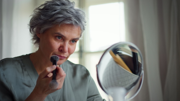 Attractive mature woman at home, sitting at cosmetic table, putting in powder with brush, observing herself in mirror. Detail shot.