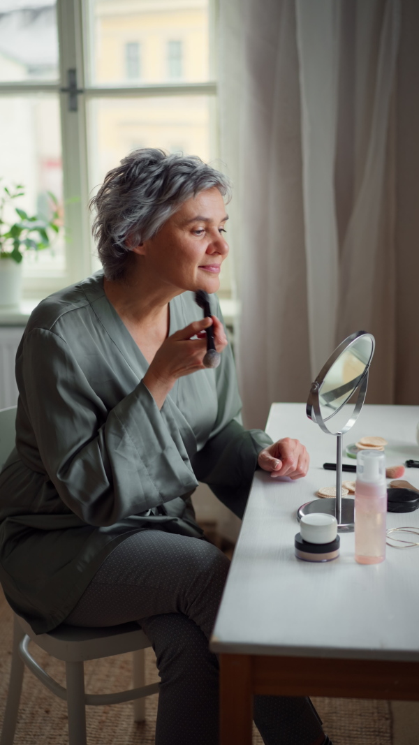 Attractive mature woman sitting at cosmetic table, looking into mirror, putting on make up. Body positivity and aging gracefully concept.