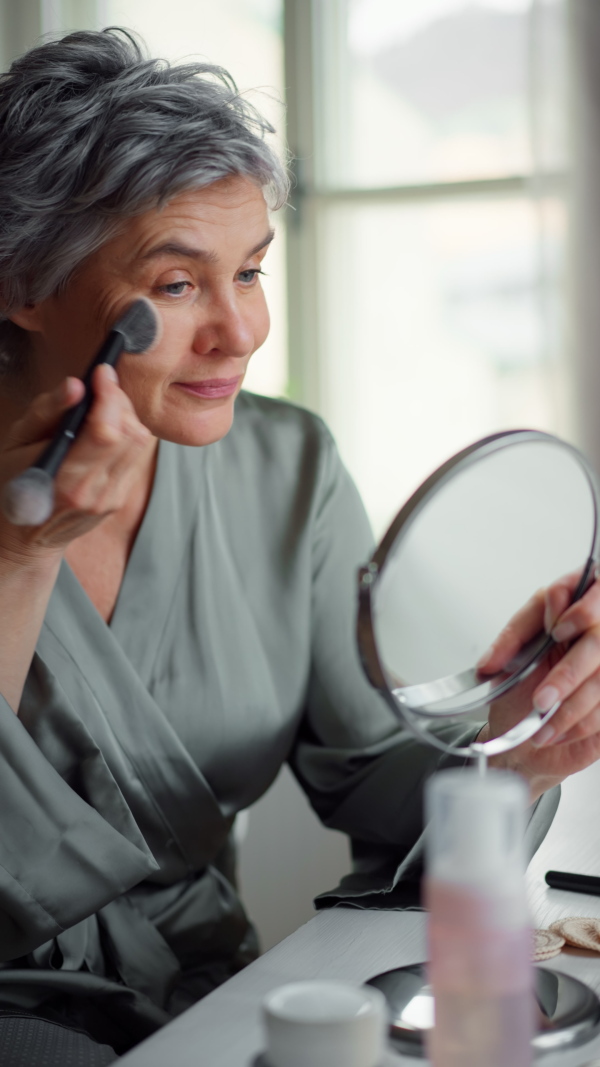 Close up portrait of beautiful mature woman applying make up powder with brush, looking in mirror, smiling. Vertical shot.