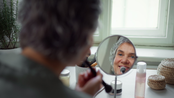 Close up of mature woman looking at reflection in mirror, putting on blusher. Body positivity and aging gracefully concept.