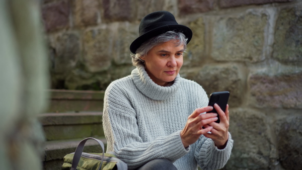 Portrait of stylish attractive mature woman sitting on stairs in town, using phone and smiling into camera. Camera raid, close up.