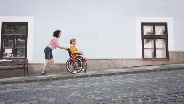 Young woman assisatant pushing older woman on wheelchair on street pavement up the hill. Side view full shot.