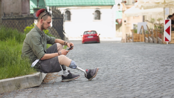 Young authentic disabled man with leg prosthetis sitting on pavement, using mobile phone, looking in camera, smiling.