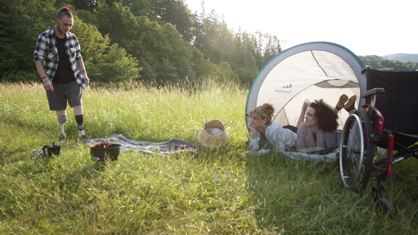 Disabled woman and young man with leg prosthetis enjoying barbeque in nature with friend, lying in tent.