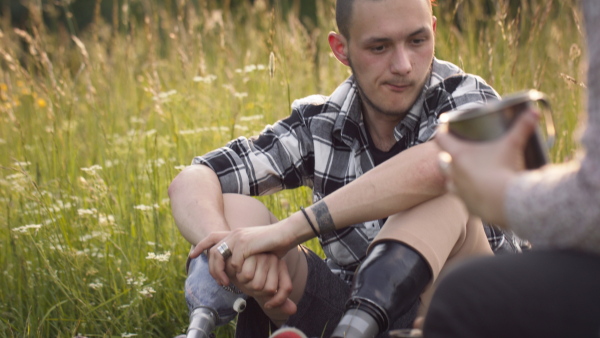 Disabled teenager with leg prosthetis sitting on sunlit meadow, looking around. while having a picnic with friends.