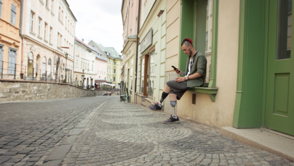 Disabled young man with leg prosthetis, sitting in hostorical town street, looking at mobile phone, smoking.