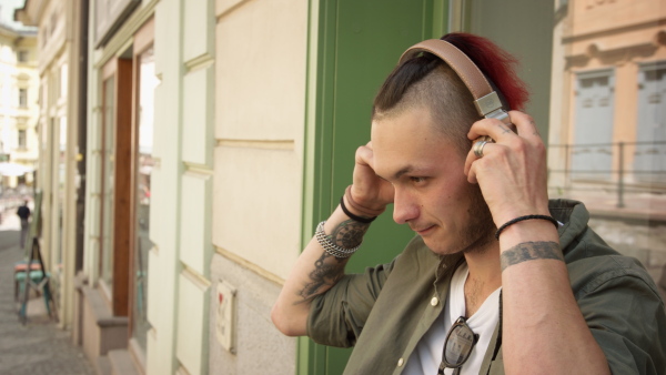 Young man with tattoo and original haircut putting on headphones, listening to music on street.