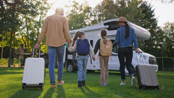 A rear view of young family with suitcases going to caravan outdoors at park.