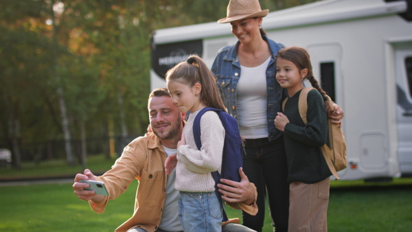 A happy young family with two children taking selfie with caravan at background outdoors.