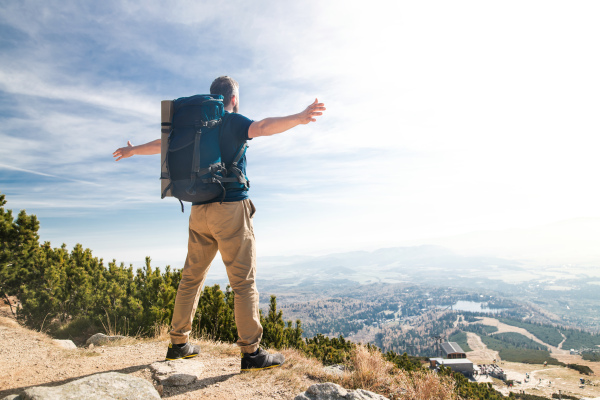 Rear view of man with backpack hiking in mountains in summer, resting with arms stretched.