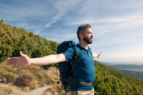 Mature man with backpack hiking in mountains in summer, resting with arms stretched.