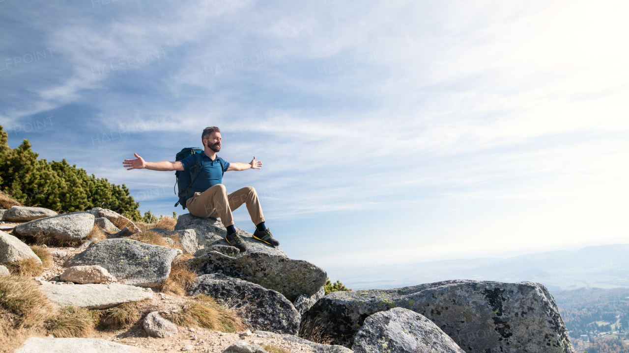 Mature man with backpack hiking in mountains in summer, resting with arms stretched.