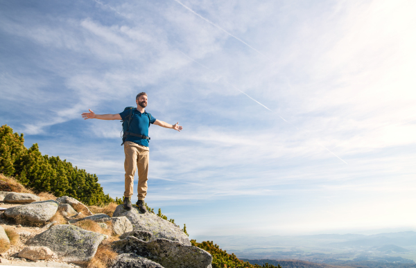 Mature man with backpack hiking in mountains in summer, resting with arms stretched.