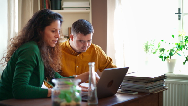 A young man with Down syndrome with his tutor studying and using laptop indoors at home, homeschooling.