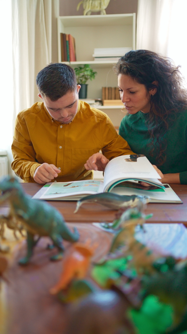 A vertical footage of mid adult woman mentoring young man with Down syndrome, reading book about dinosaurs indoors at home.