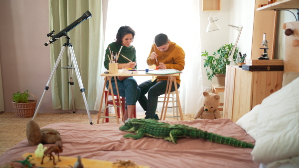 A young man with Down syndrome with his tutor painting pictures indoors at home, homeschooling.