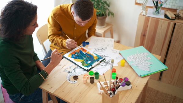 A young man with Down syndrome with his tutor painting pictures indoors at home, homeschooling.