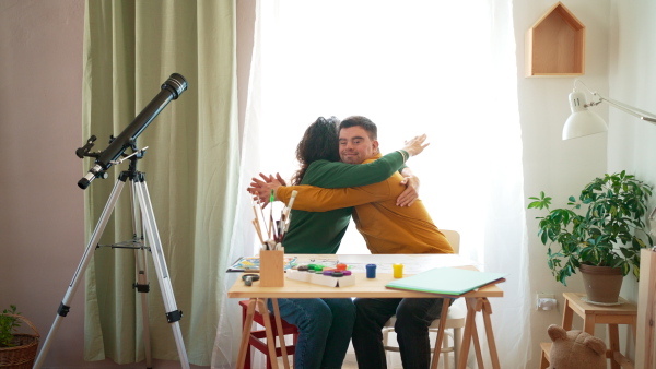 A young man with Down syndrome hugging his tutor when painting pictures indoors at home, homeschooling.