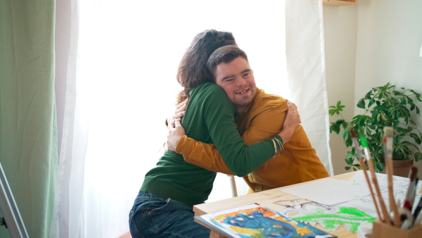 A young man with Down syndrome hugging his tutor when painting pictures indoors at home, homeschooling.