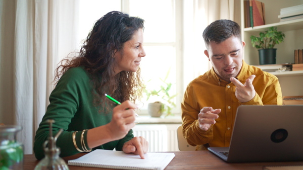 A young man with Down syndrome with his tutor studying and using laptop indoors at home, homeschooling.