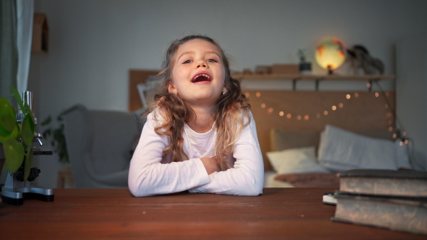 A happy cute little girl sitting at desk at home and looking at camera.