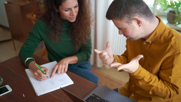 A young man with Down syndrome with his tutor studying and using laptop indoors at home, homeschooling.