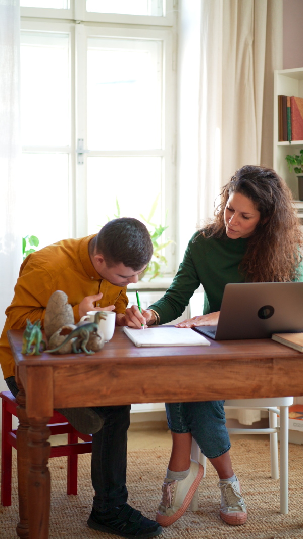 A vertical footage of young man with Down syndrome with his tutor studying and using laptop indoors at home, homeschooling.