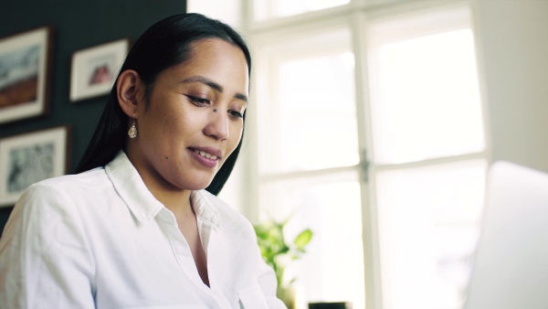 A young asian woman with laptop sitting at the desk indoors at home, typing.