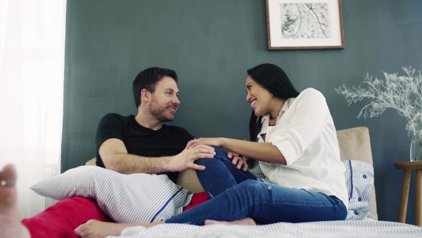 A young affectionate couple sitting on bed indoors at home, talking.