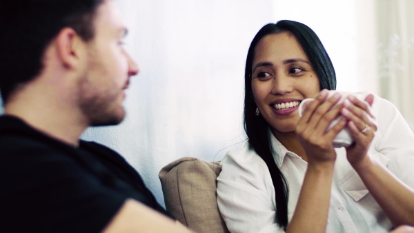 A close-up of young affectionate couple with coffee sitting indoors at home, talking.