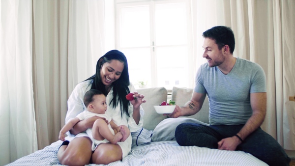 A young happy couple with a baby indoors on bed, eating strawberries.